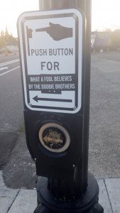 A picture of a sign at a crosswalk where someone put a sticker over the "cross" section of "push button for crossing" so it now reads "push button for what a fool believes by the doobie brothers" & there's a self help radio logo on the button you push