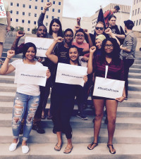 A group of youth of color stand on outdoor stairs with TL Lewis. They are Deaf, DeafBlind, DeafDisabled, and Hard of Hearing young people at the national prison strike rally.