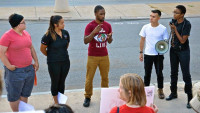 on the sidewalk at the national prison strike rally, a group of people of color stand in a circle. TL speaks into a loud speaker while someone ASL interprets TL. The person interpreting has a shirt with "ASL Live" and an image of an eye.