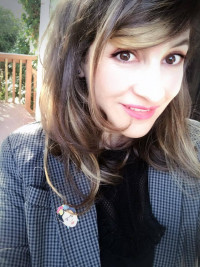 Sara outside on a sunny day smiling for the camera. She has blonde highlights, a herringbone jacket over black ruffly blouse, and a Frida Kahlo lapel pin.